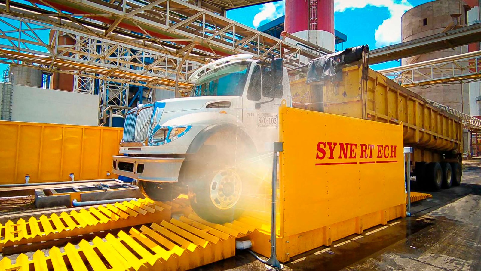 Camión grande blanco y amarillo pasando por un sistema de lavado de ruedas activo, rociando agua para limpiar la suciedad y escombros. En un sitio industrial o de construcción con silos al fondo, el sistema ayuda a mantener limpias las vías públicas. - ver detalles