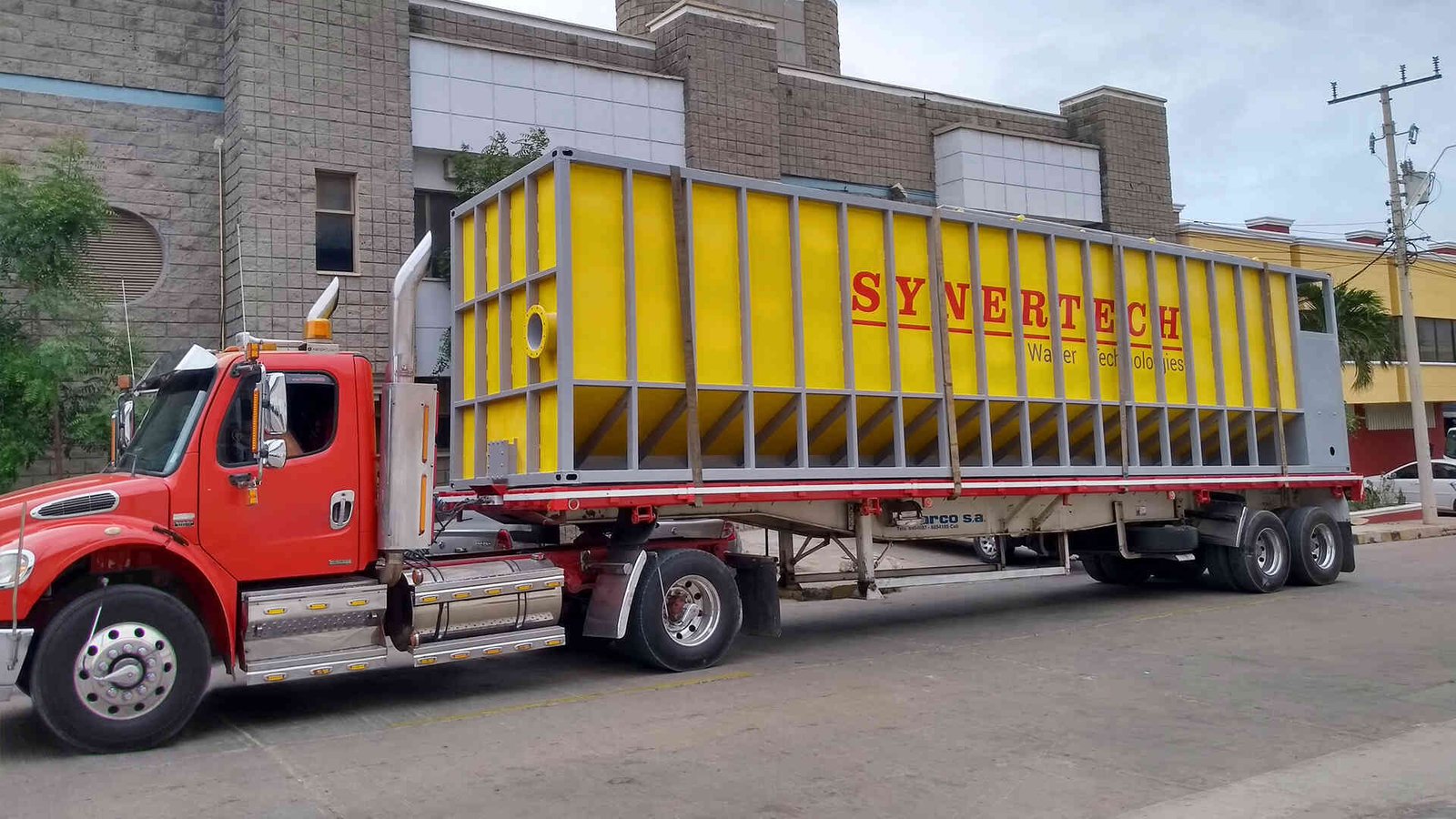 Camión rojo de plataforma larga transportando un gran decantador amarillo de la marca 'SYVERTECH' para el tratamiento de aguas residuales complejas, estacionado en una calle urbana con edificios y cielo despejado al fondo. - ver detalles