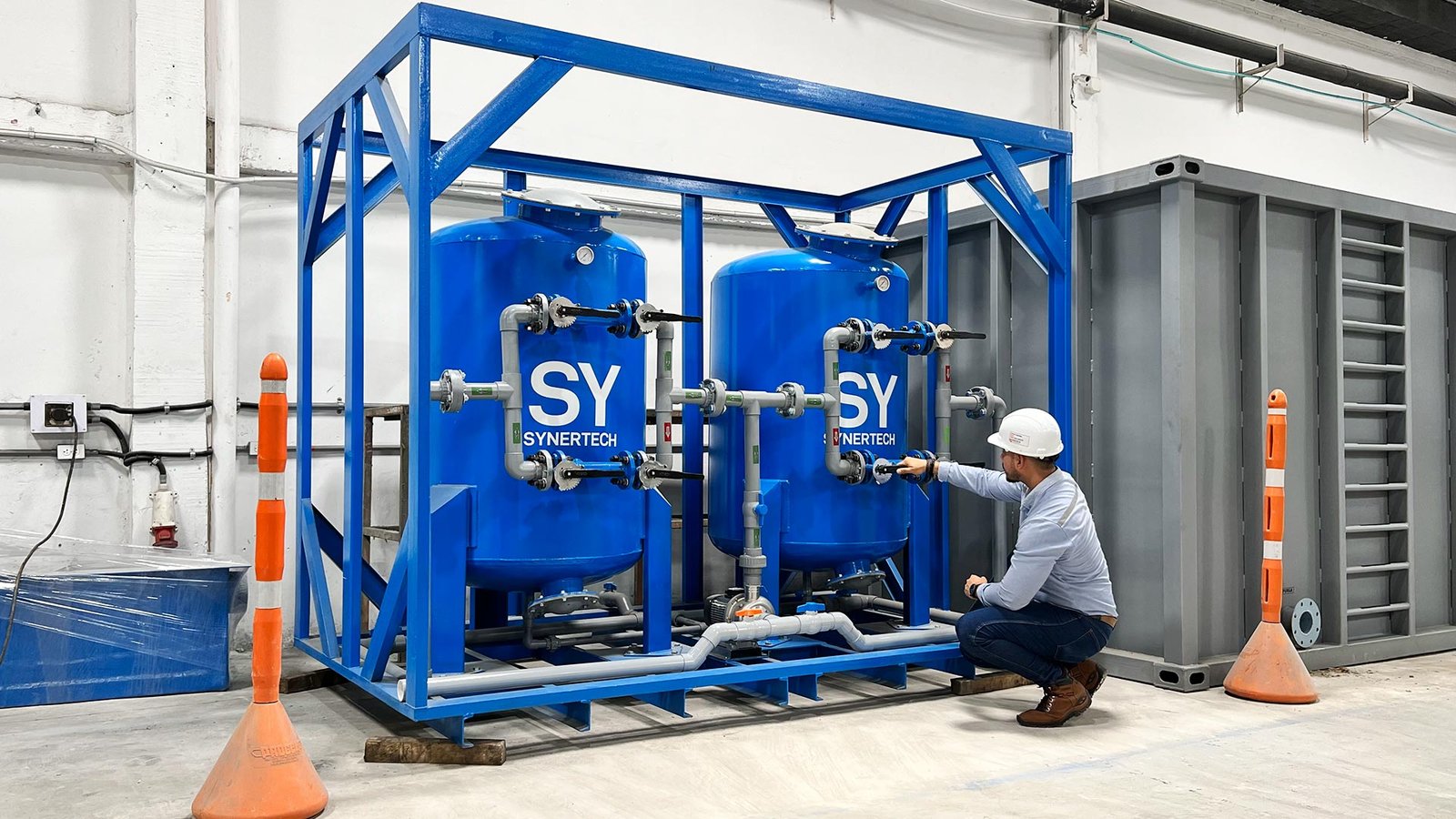 Técnico inspeccionando una estación de filtración con dos filtros azules de la marca Synertech, conectados a un sistema de tuberías. El equipo está instalado en un entorno industrial con estructuras y elementos de seguridad visibles.