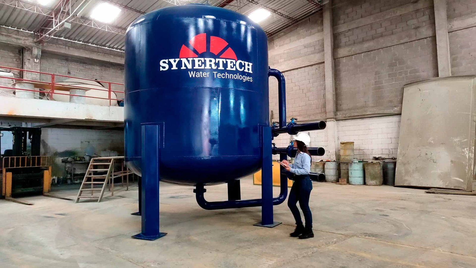 Técnico inspeccionando un filtro multimedia industrial azul de gran tamaño, marca Synertech Water Technologies, en un entorno de planta o fábrica. El equipo se encuentra dentro de un edificio con estructura de concreto y tuberías conectadas al filtro. - ver filtros